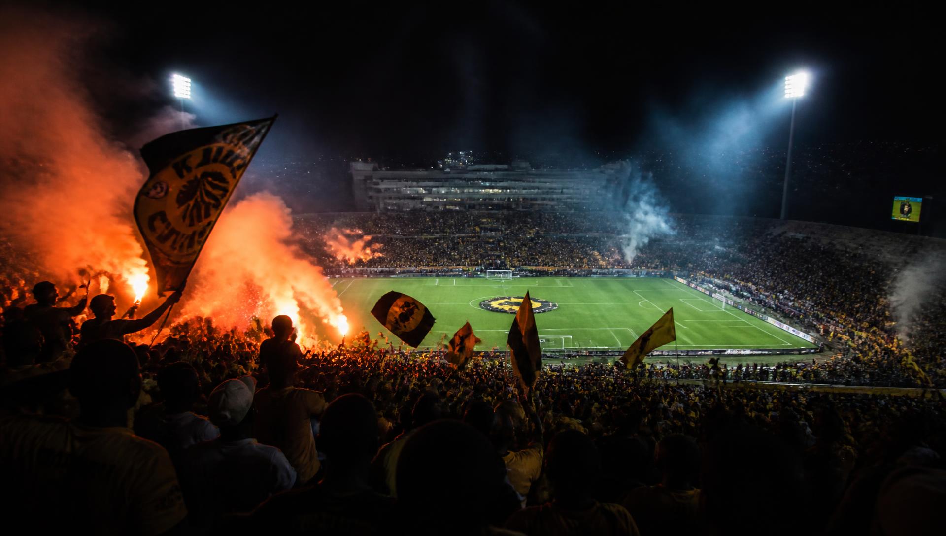 Packed African football stadium at night with fans, flags and floodlights